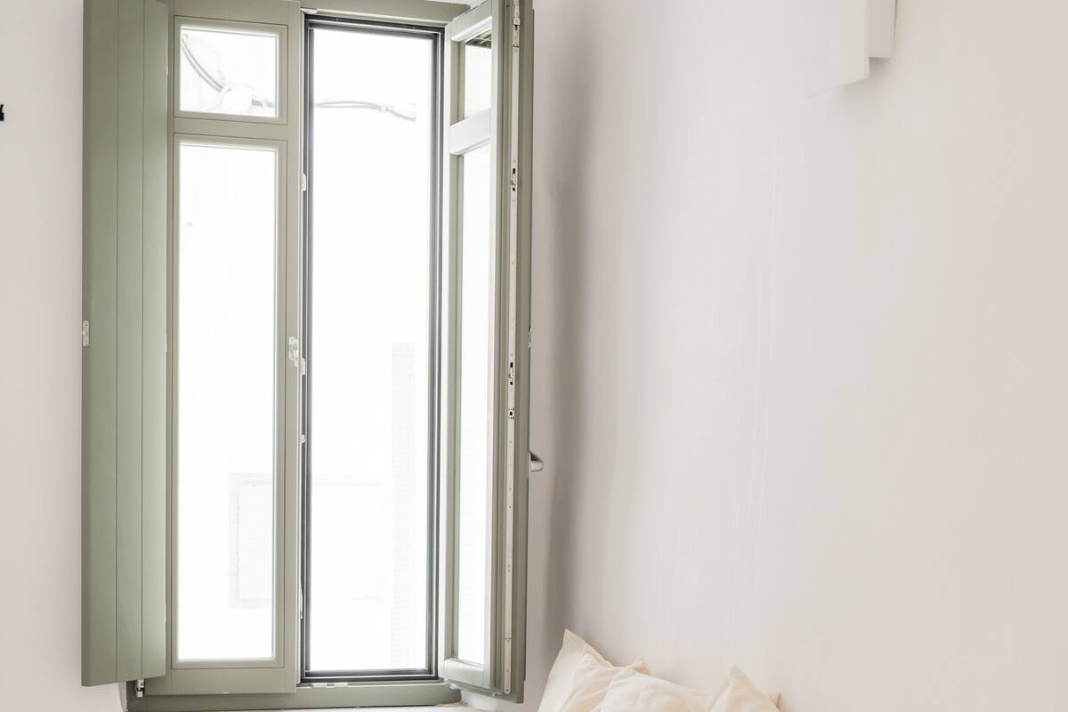 A minimalist bedroom with a bed, a window with green shutters, a small plant, and a bedside table with a book.