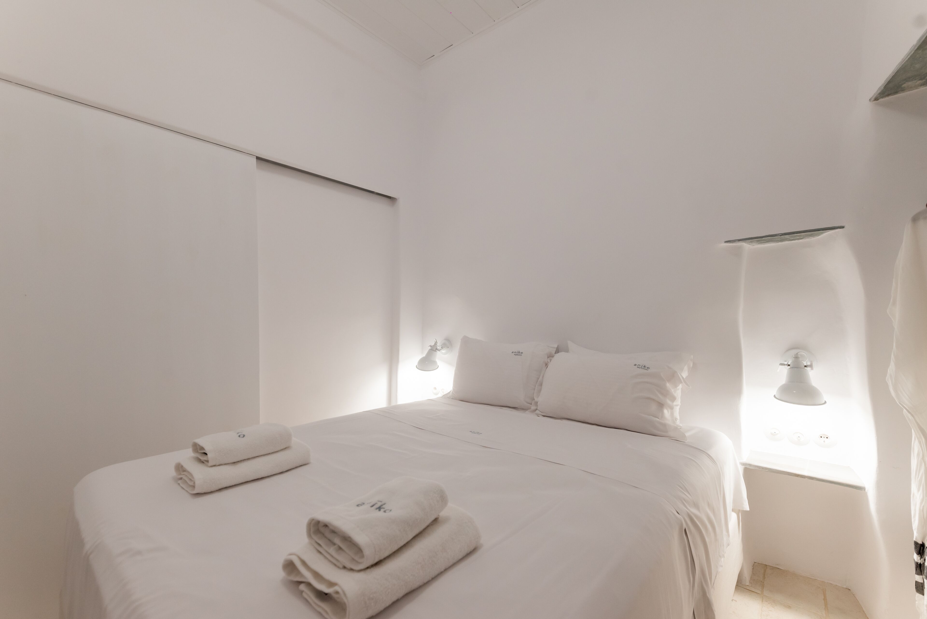 A minimalist white room with a bed, two bedside lamps, and a window with a view.