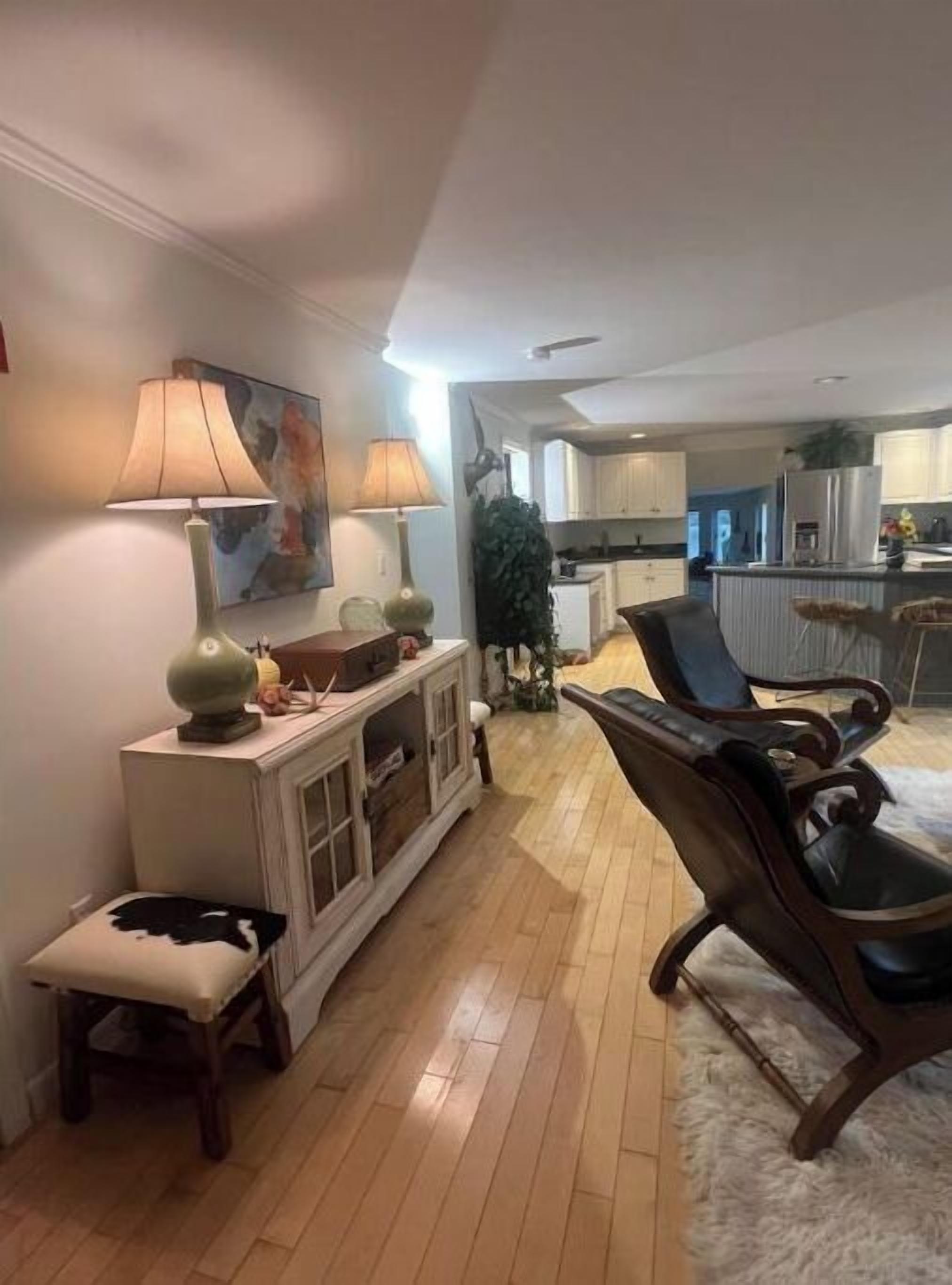 Een ruime woonkamer met een houten vloer, een grote fauteuil, een console tafel met lampen, en een schilderij aan de muur.