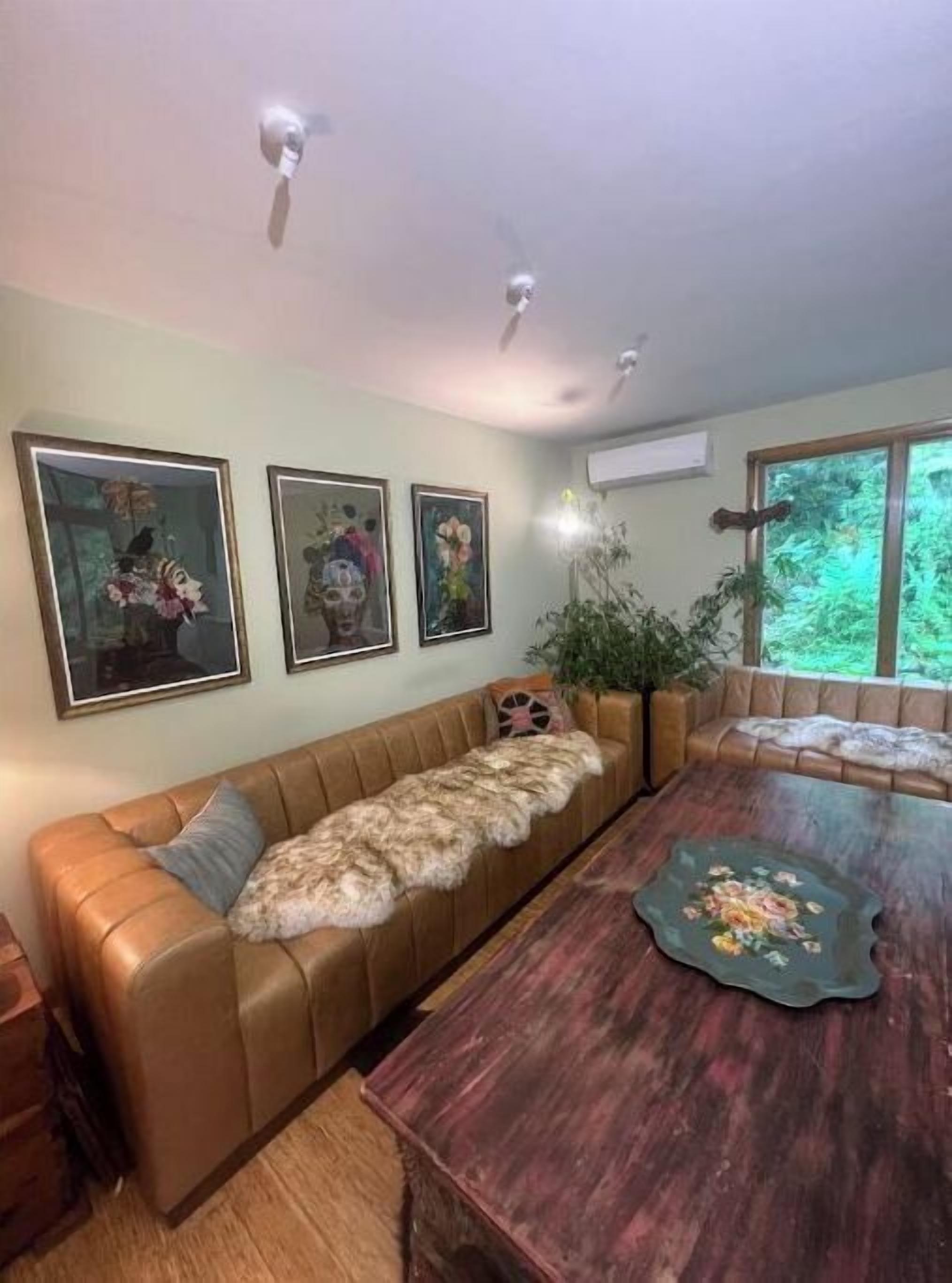 A living room with a brown leather sofa, a wooden coffee table with a floral centerpiece, and three framed artworks on the wall.