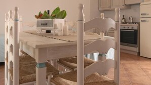 Uma mesa de jantar com acabamento branco, rodeada por quatro cadeiras com almofadas trançadas nos assentos. Uma pequena planta e um recipiente branco estão sobre a mesa.