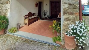 Edifici de pedra amb un porxo de rajola, una banca de fusta i plantes en testos.