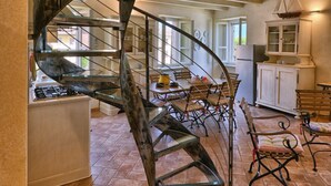 Una escalera de caracol con diseño rústico que conduce a un piso superior. Abajo se ve la zona de la cocina, con una estufa a gas y una mesa de comedor con sillas.