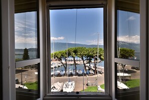 A view through a window of a marina with boats docked, trees, and a clear sky. - Al Porto Vecchio by Theater Home - with parking space and marvellous lake view (Gargnano)