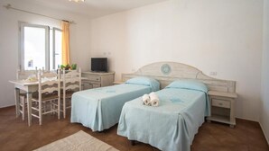 A bedroom with two beds, a desk, a chair, a TV, and a window with curtains.