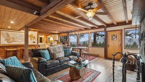 A spacious living room with a wooden ceiling, leather sofas, a glass coffee table, and large windows offering a view of the outdoors. - Big Mountain Treehouse!  Beautiful cabin home w/ SPECTACULAR views of Lake (Whitefish)