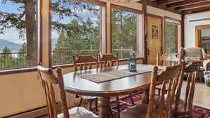 A wooden dining table with six chairs, large windows with a view of trees and mountains, and a chandelier. - Big Mountain Treehouse!  Beautiful cabin home w/ SPECTACULAR views of Lake (Whitefish)