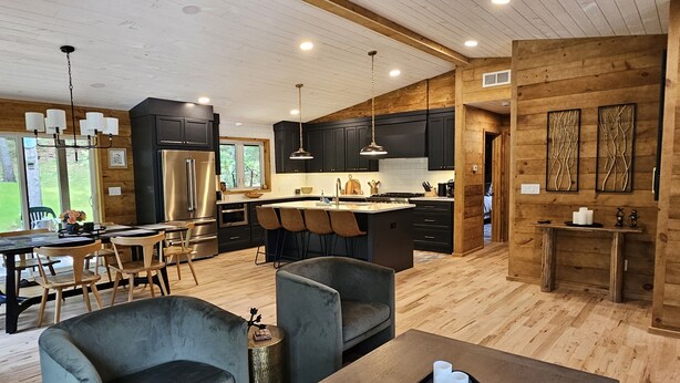 A modern kitchen with dark cabinets, a central island, and a dining area with a table and chairs. - Cabin with hot tub, heated swimming pool and acres of backyard hiking  (Adams)