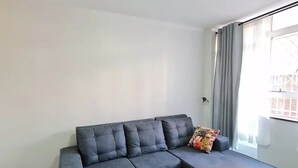 A modern living room with a sectional sofa, a patterned rug, and a large window with curtains. - Heritage Self Catering Apartment<br>Home from Home (Harare)