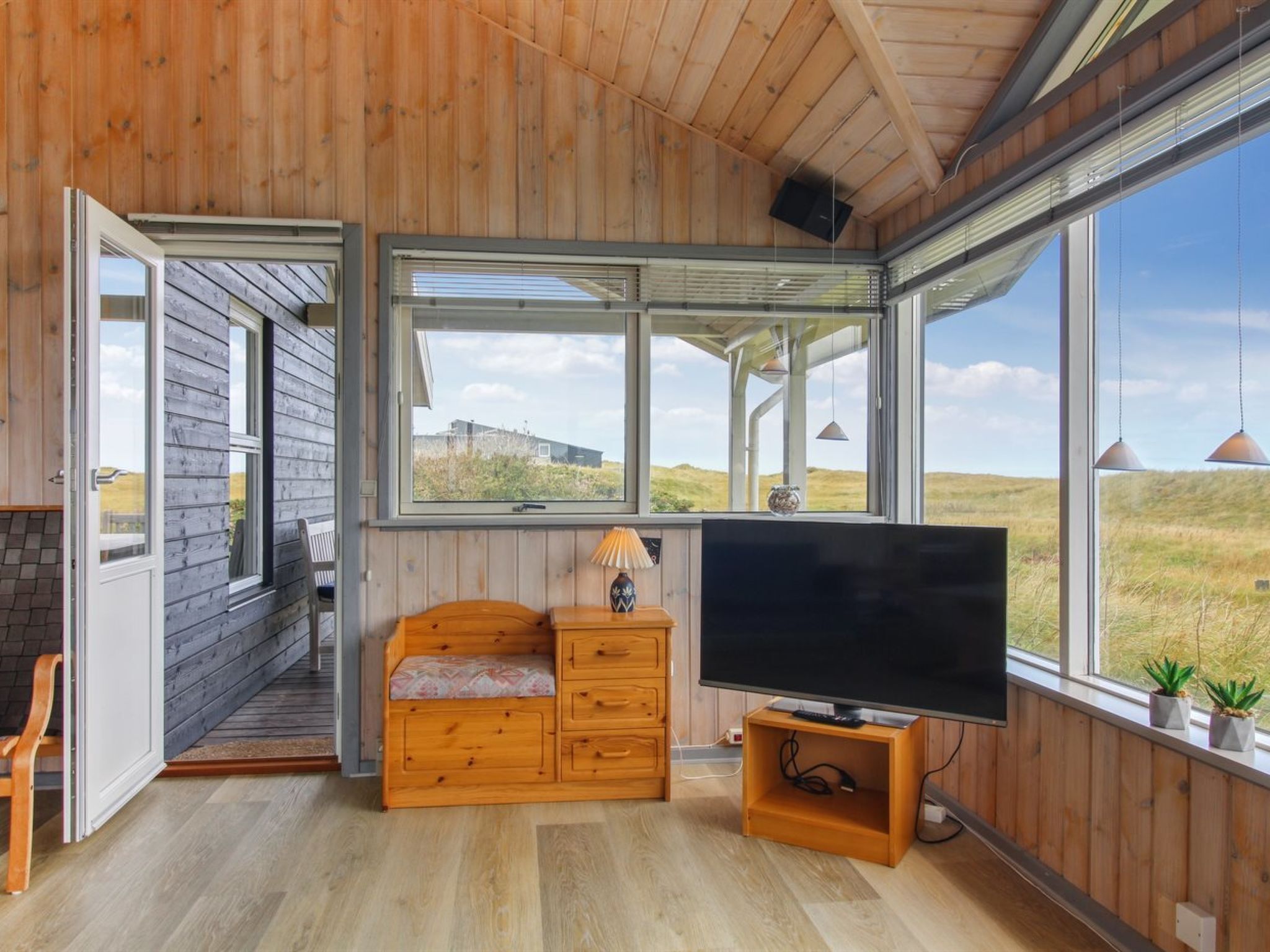 Une pièce aux murs et au plancher en bois, une grande fenêtre donnant sur la campagne, un banc en bois, un téléviseur posé sur un meuble en bois et une petite table avec une lampe.