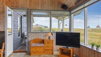 Une pièce aux murs et au plancher en bois, une grande fenêtre donnant sur la campagne, un banc en bois, un téléviseur posé sur un meuble en bois et une petite table avec une lampe.