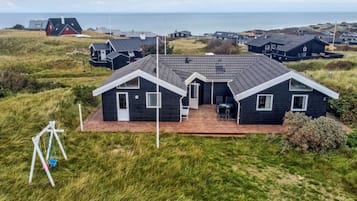 Une maison située sur la côte, avec une façade sombre, une terrasse dotée de mobilier d’extérieur et une vue sur la mer.