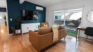 A modern living room with a brown leather sofa, a flat-screen TV mounted on a teal wall, and a balcony with potted plants. - ☞Kid Paradise☞5miles->Disneyland☞1 Parking☞W/D☞AC (Santa Ana)