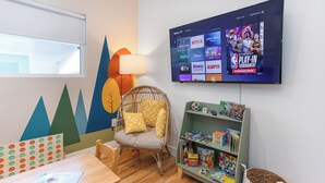 A cozy room with a TV displaying a sports event, a wicker chair, a bookshelf with children's books, and a wooden table. - ☞Kid Paradise☞5miles->Disneyland☞1 Parking☞W/D☞AC (Santa Ana)