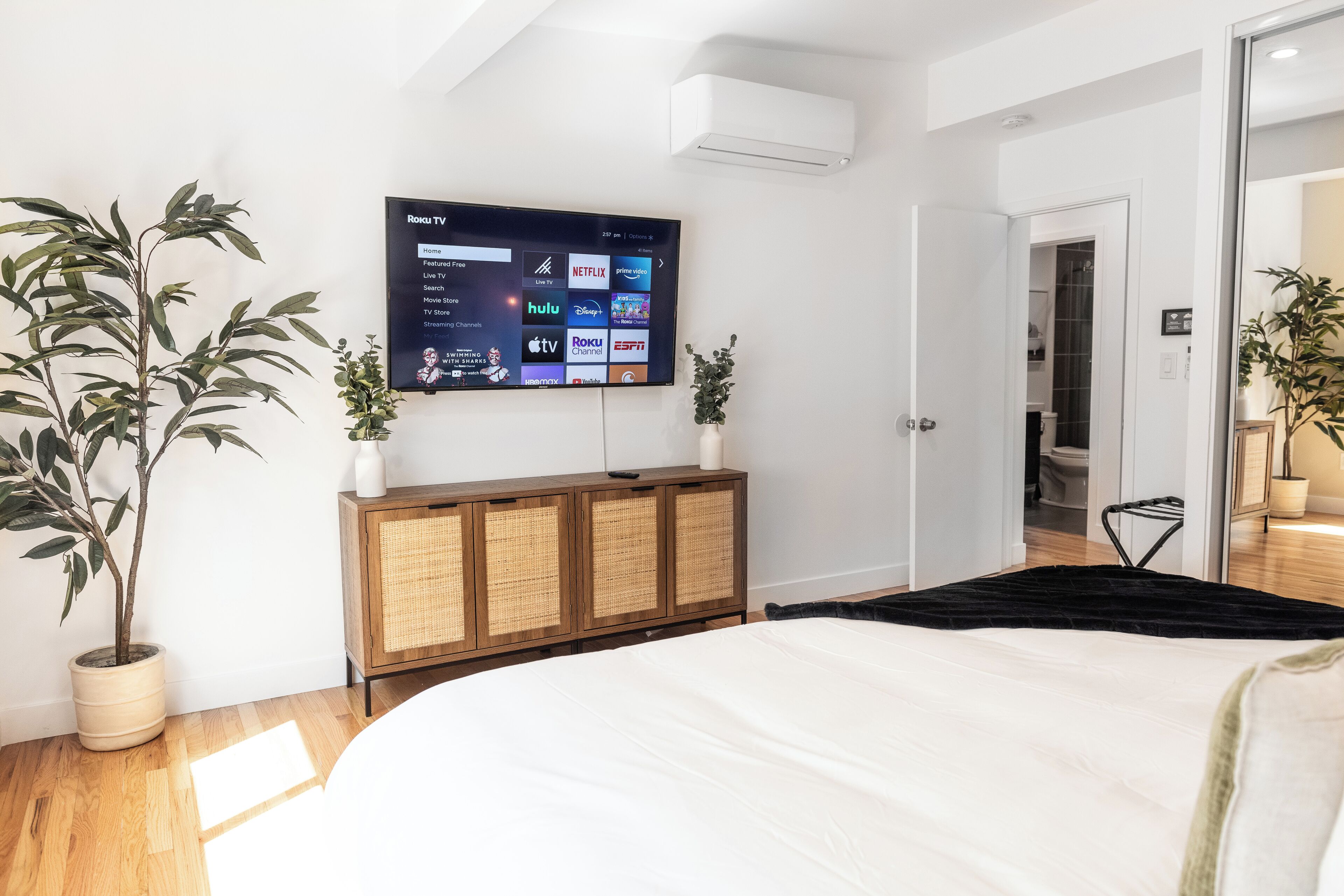 A modern bedroom with a large bed, a mounted TV displaying streaming services, a wooden cabinet, and two potted plants.