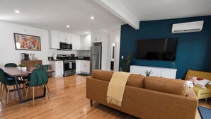 A modern living room with a brown sofa, a dining table with chairs, a kitchen area with white cabinets, a flat-screen TV mounted on a blue wall, and an air conditioning unit. - ☞Kid Paradise☞5miles->Disneyland☞1 Parking☞W/D☞AC (Santa Ana)