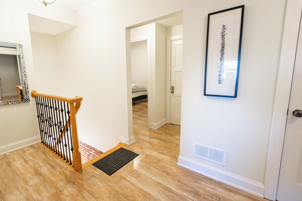 A hallway with a wooden staircase, a framed picture on the wall, and a door at the end. - Spacious 3 Bedroom Apartment in Heart of Niagara (Thorold)