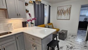 A modern kitchen with grey cabinets, a white countertop, and a marble floor. There is a yellow sofa and a black stool in the background.