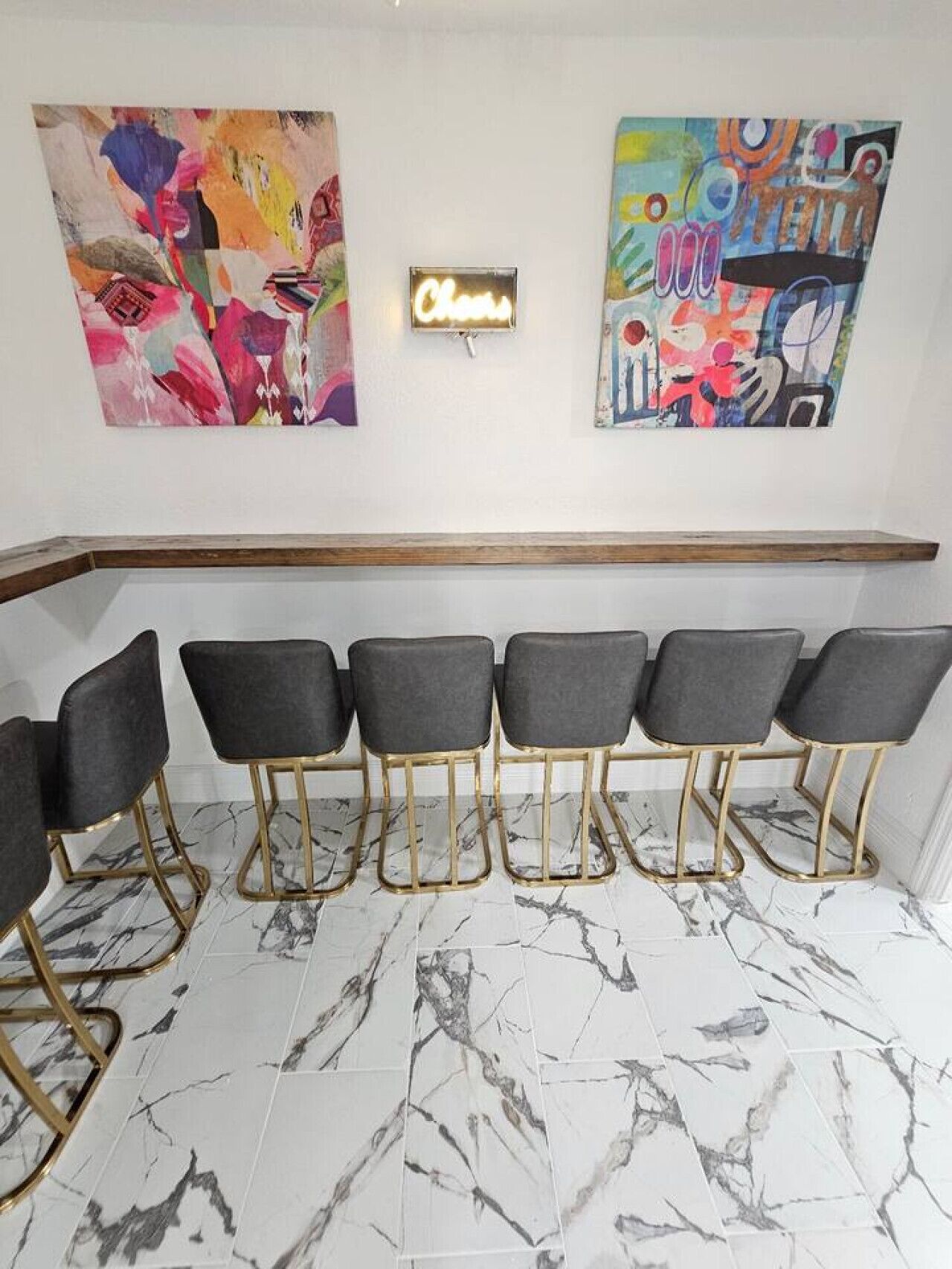 Een bar met zes zwarte barkrukken met gouden poten, een marmeren vloer en twee abstracte schilderijen aan de muur.