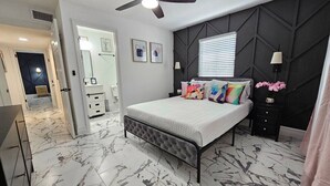 Un dormitorio con cama, ventilador de techo, mesita de noche, cómoda y ventana con persianas.
