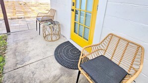 Eine überdachte Terrasse mit zwei Korbstühlen und einem kleinen Tisch, einer gelben Tür und einer weißen Wand.