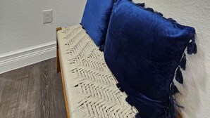 Ein Zimmer mit weißer Wand, einem gerahmten Bild, einer hängenden Pflanze, einem Fenster mit Jalousien und einem Bett mit blauen Kissen.