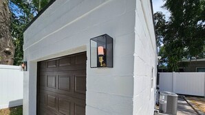Eine Garage mit brauner Tür, weißer Außenwand und einer kleinen, schwarz gerahmten Wandleuchte.