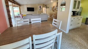 Un comedor con una mesa de madera y sillas blancas, un sofá y una cocina al fondo.