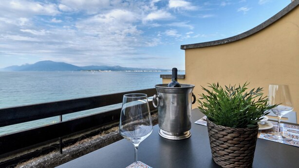 A table with a wine glass, a metal pot, and a potted plant on a balcony overlooking a body of water and distant mountains. - Lydie apt. exclusive holigarda (Desenzano del Garda)