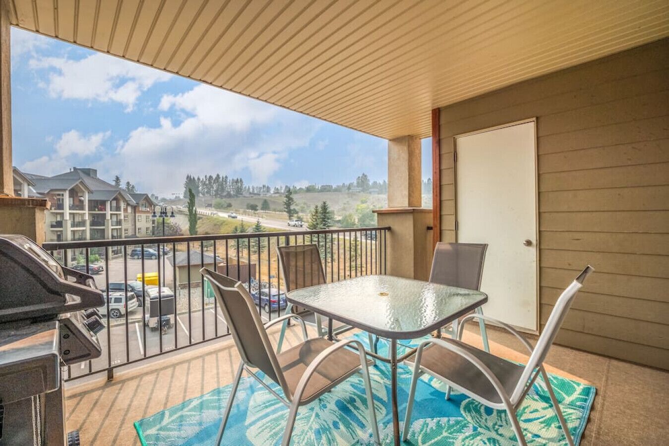 A balcony with a glass table and chairs, a barbecue grill, and a view of a residential area.
