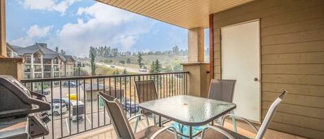 A balcony with a glass table and chairs, a barbecue grill, and a view of a residential area.