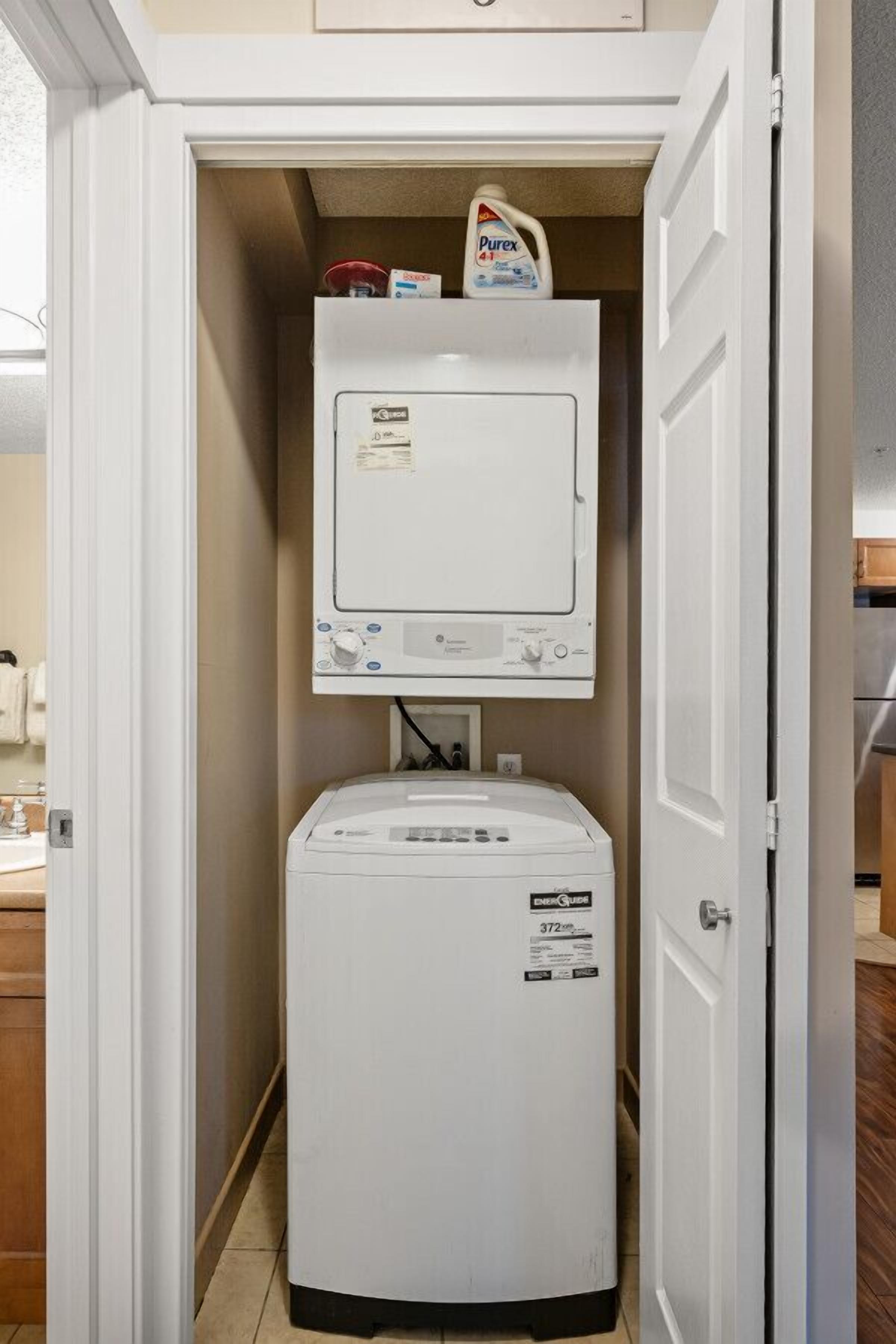 Een kleine wasruimte met een voorlader wasmachine en droger, een klein plankje met een fles Purex wasmiddel, en een magnetron boven de droger.