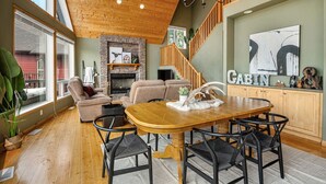 A spacious living area with a wooden dining table, cushioned seating, a fireplace, and a staircase leading to an upper level. - Cedar & Sage | Hot Tub | A-Frame | Family Friendly (Radium Hot Springs)
