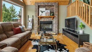 A cozy living room with a stone fireplace, a sofa, a coffee table, and a TV. - Cedar & Sage | Hot Tub | A-Frame | Family Friendly (Radium Hot Springs)