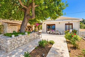 Une maison moderne dotée d’un patio en pierre, d’un espace salon extérieur et d’un grand arbre qui offre de l’ombre.
