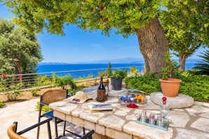 A stone table with a bottle, glasses, and a bowl of fruit, overlooking a scenic coastal view. - Villa Ariadne - Five Bedroom Villa, Sleeps 10 (Apergatika)