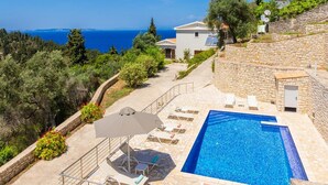 Piscina con sillas de descanso y una sombrilla, rodeada de muros de piedra y vegetación, con vista al mar.
