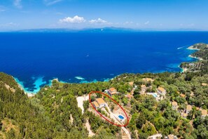 An aerial view of a coastal property with a swimming pool, surrounded by dense greenery and a clear view of the sea.
