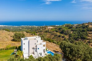 Une villa moderne aux murs blancs, dotée d’une piscine, entourée d’une végétation luxuriante et offrant une vue sur un paysage côtier.