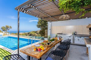 Ein Essbereich am Pool mit einem Holztisch, gedeckt mit Früchten, Blick auf das Meer sowie eine Küchenzeile mit Spüle und Schränken.