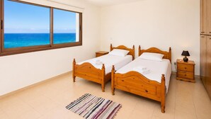 Een slaapkamer met twee houten bedden, een nachtkastje, een lamp, een raam met uitzicht op de oceaan, en een gestreept vloerkleed.