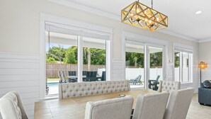Un espace repas comprenant une grande table en bois et six chaises rembourrées, de grandes fenêtres donnant sur un jardin, et un luminaire décoratif au plafond.