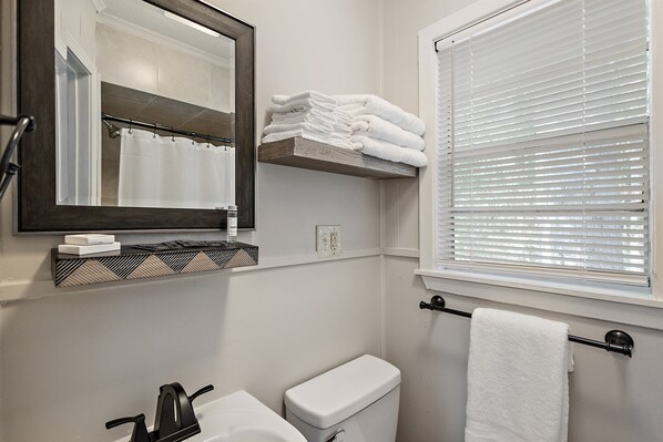 A bathroom with a toilet, sink, mirror, towel rack, and window with blinds.