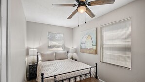 A bedroom with a bed, bedside lamps, a ceiling fan, a window with blinds, and a painting on the wall. - Cottage 5 - Smugglers Run (Ridgeland)