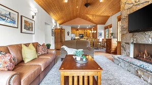 Sala de estar com lareira de pedra, teto de madeira e cozinha ao fundo.