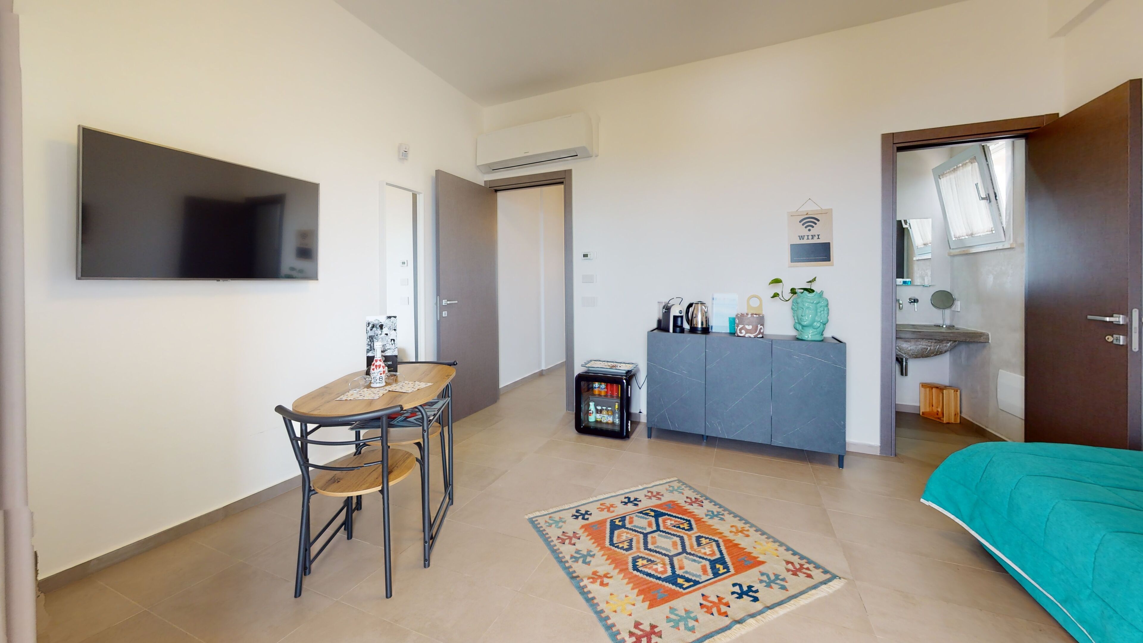 A compact hotel room with a bed, a small table with chairs, a minibar, a TV, and a door leading to another room.
