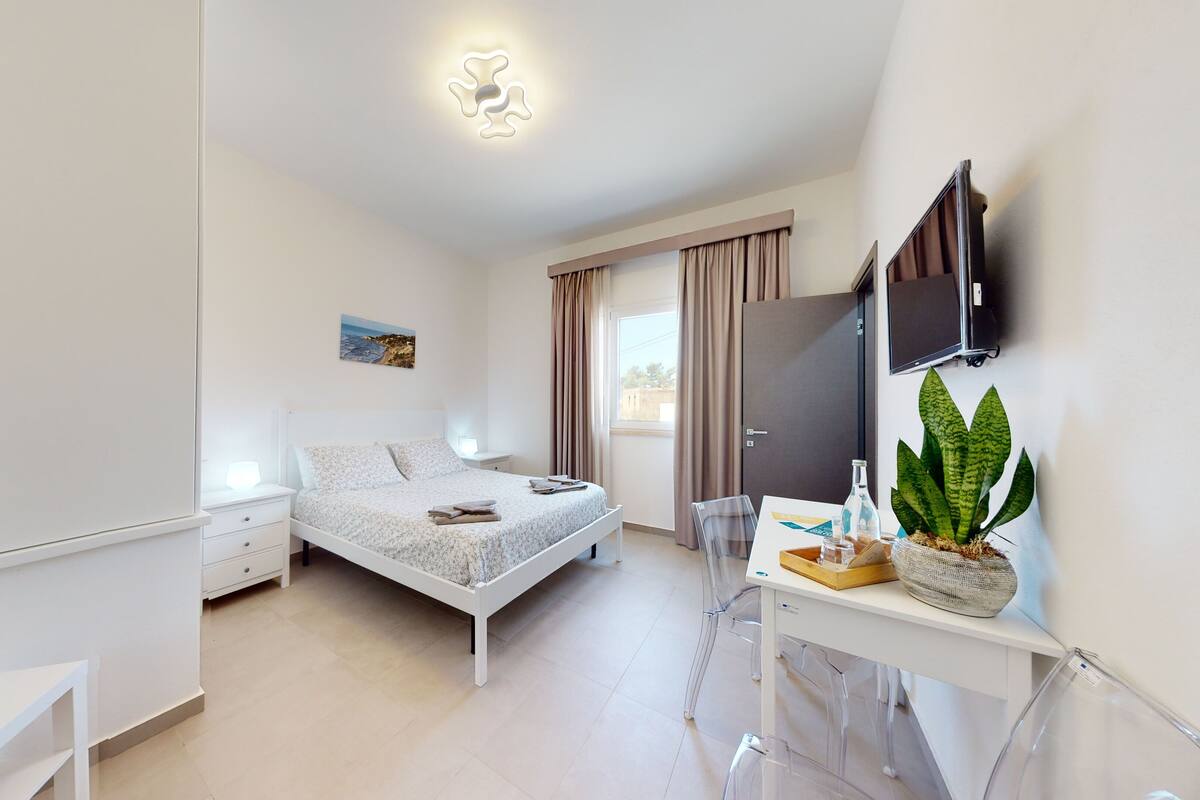 Un dormitorio moderno con cama, mesita de noche, lámpara, cortinas, televisor y una planta en maceta.