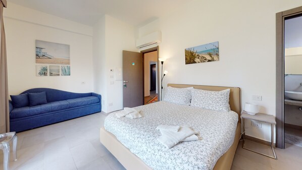 Un dormitorio con cama, sofá azul, mesita de noche y baño visible a través de una puerta abierta.