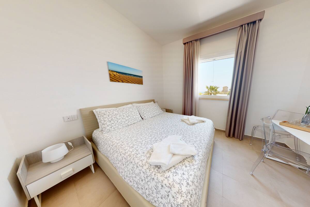 Habitación de hotel con cama, escritorio, silla, una mesita, una planta y ventana con cortinas.