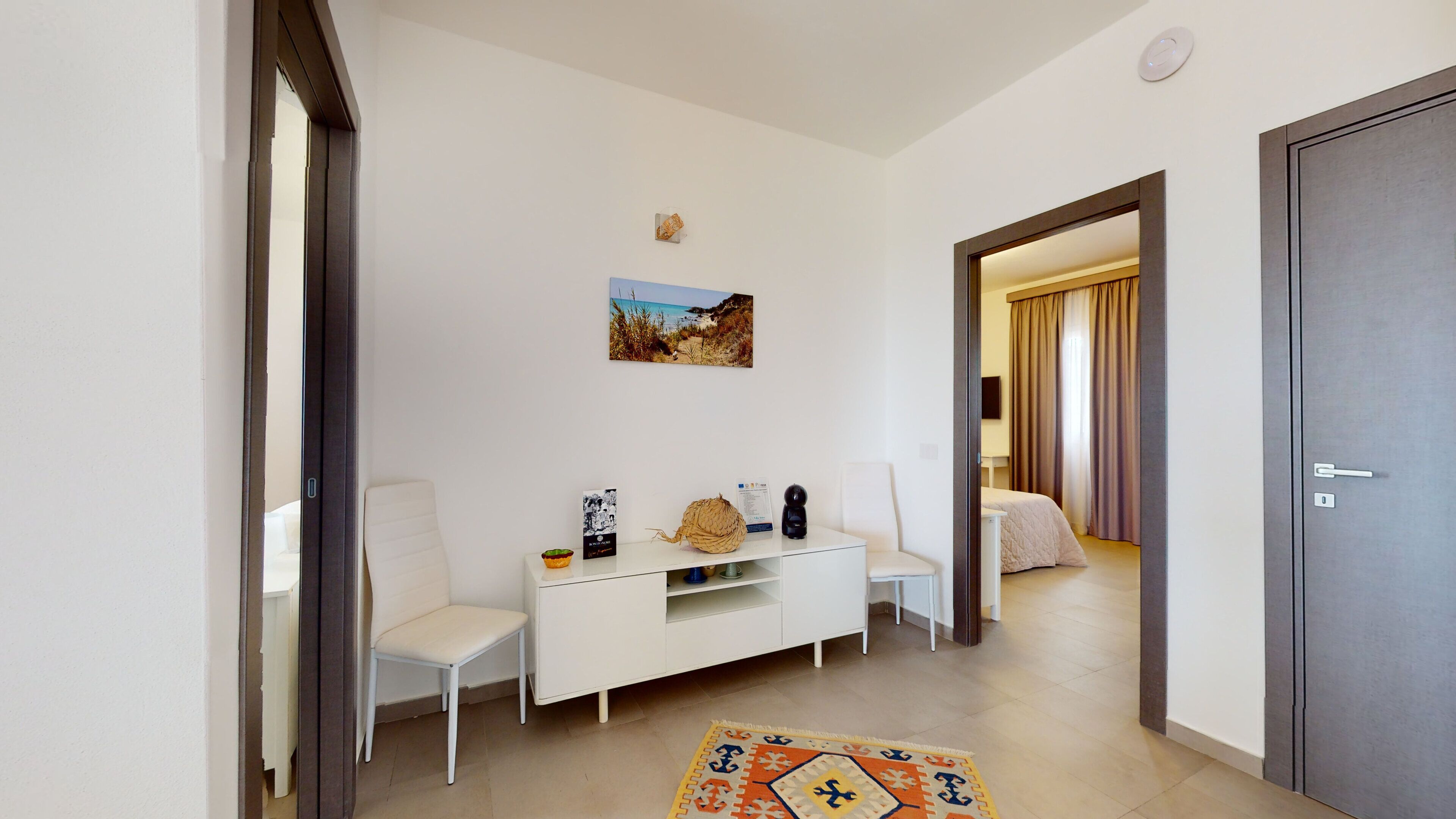 Habitación de hotel con una mesa consola blanca, una silla, una cama con cortinas, una puerta y una pequeña alfombra.